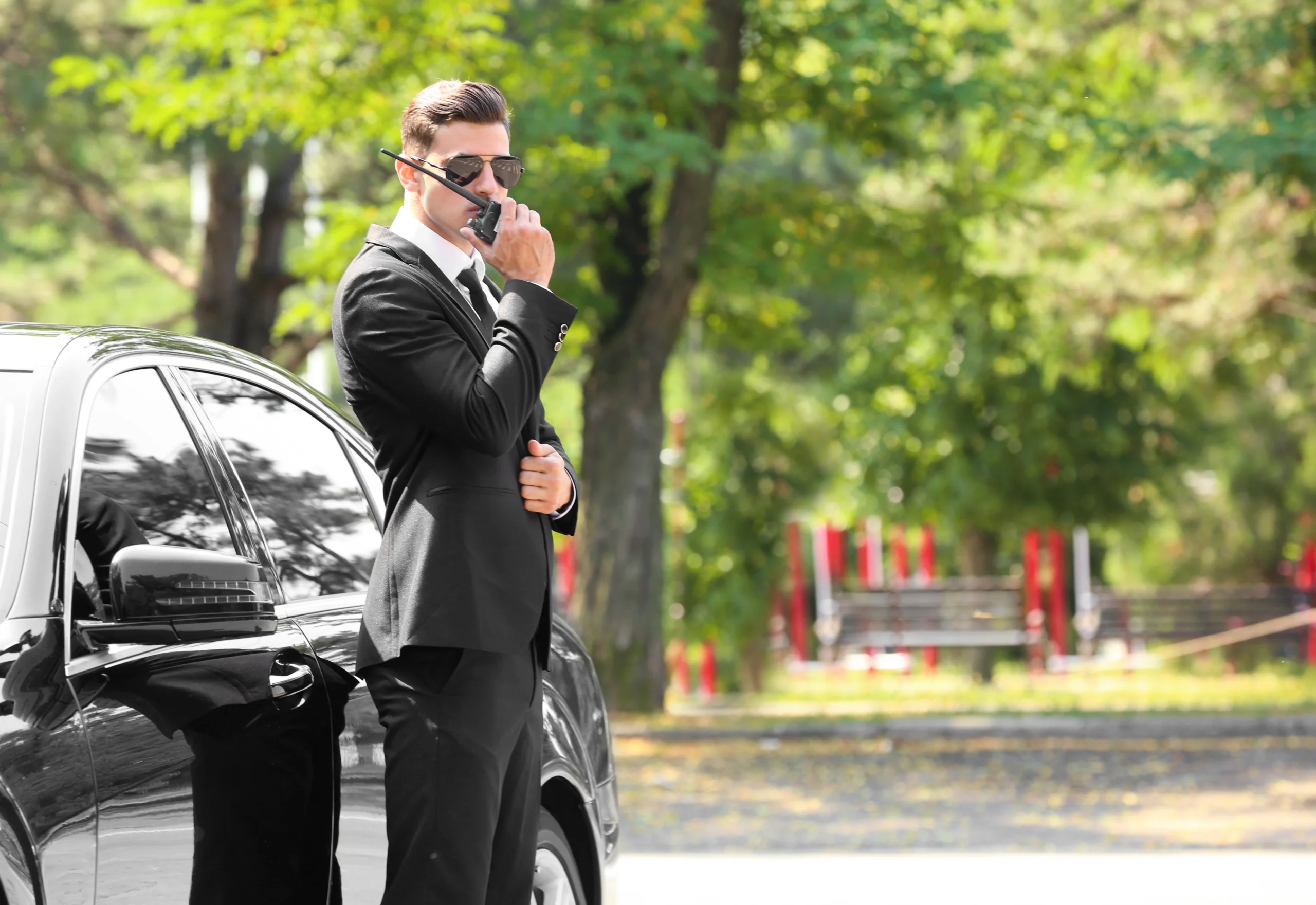 A suited bodyguard communicates via walkie-talkie beside a black vehicle.