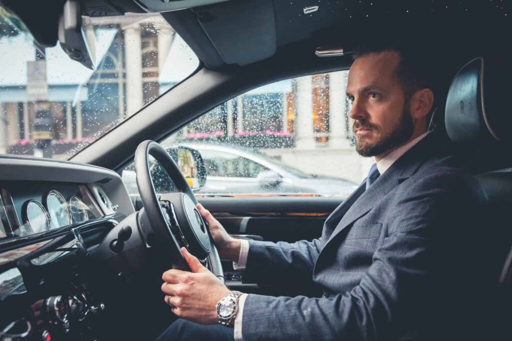 A chauffeur in a suit sits inside a high-end car during a rainy day.