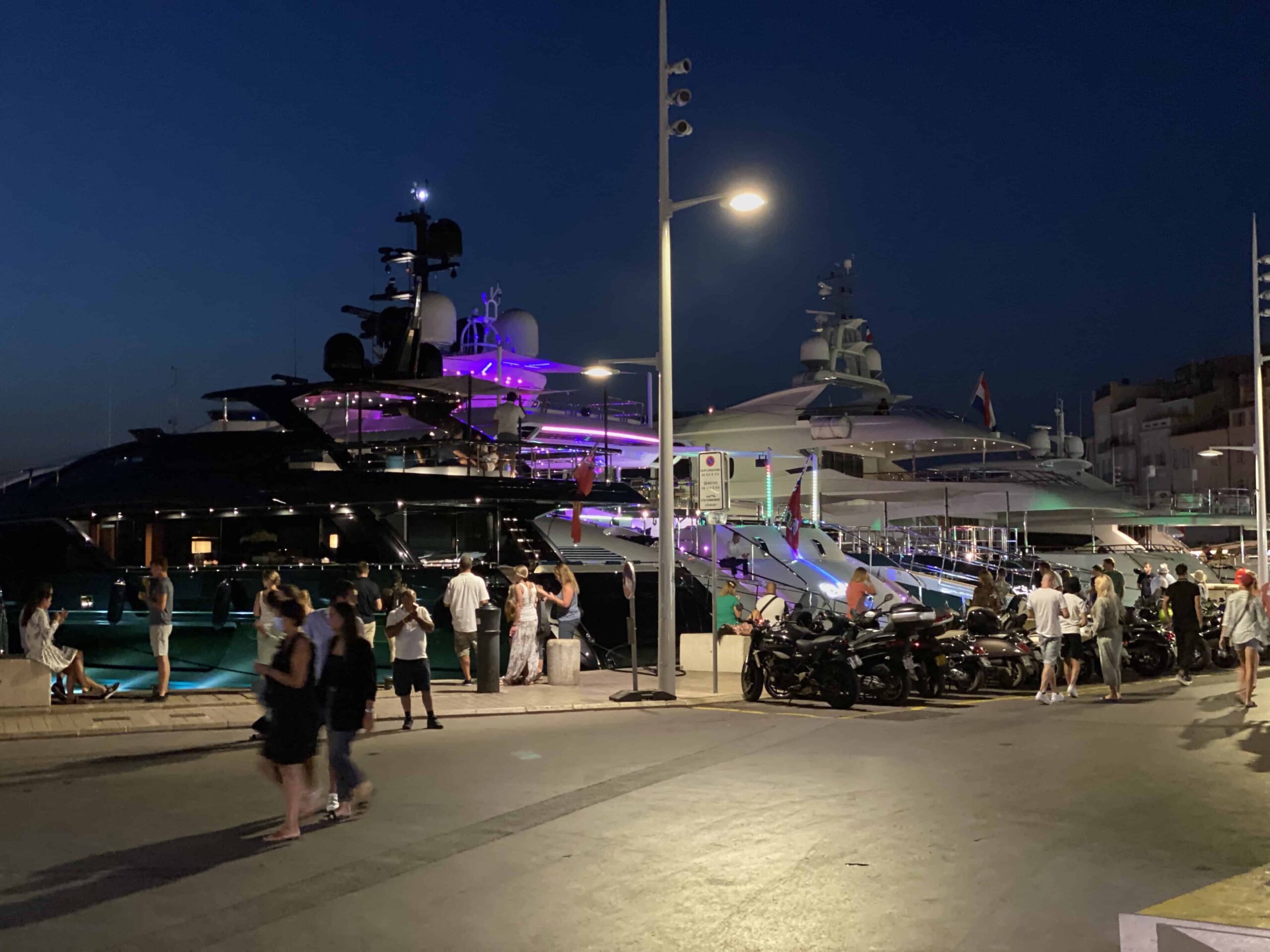 Luxury yachts docked at a harbor at night with people gathered nearby.