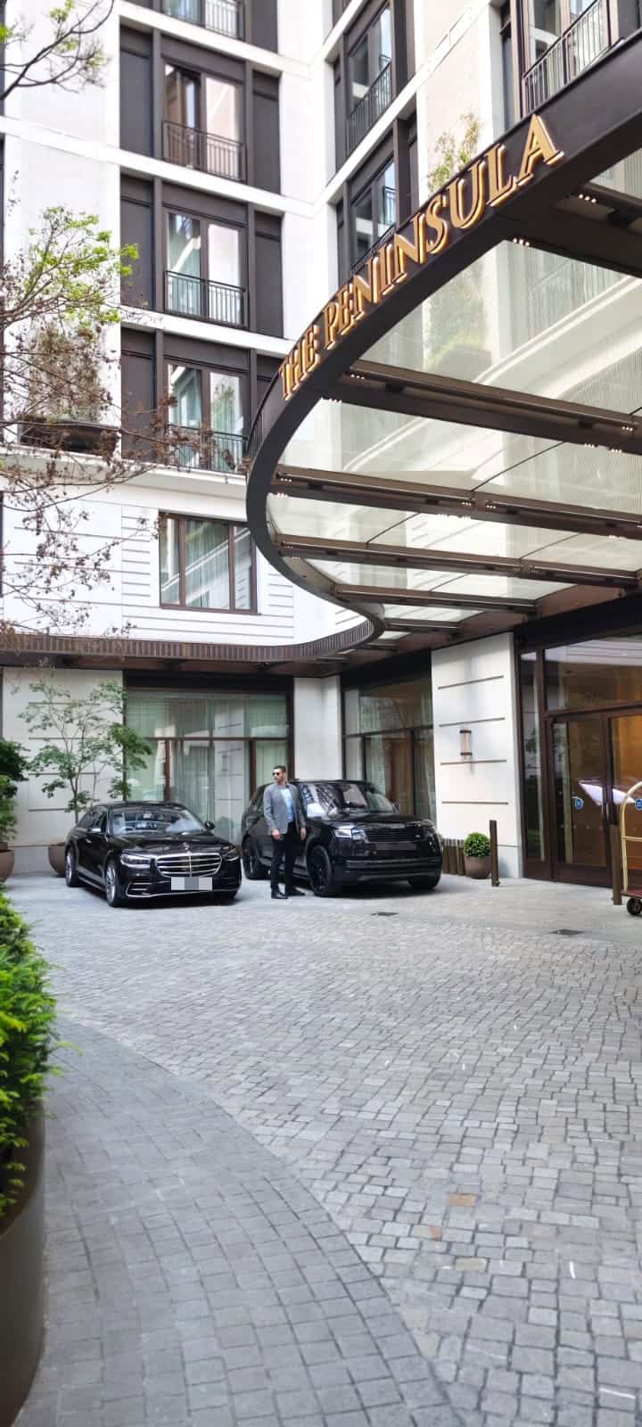 Two luxury cars parked at the entrance of The Peninsula hotel with a doorman standing outside.