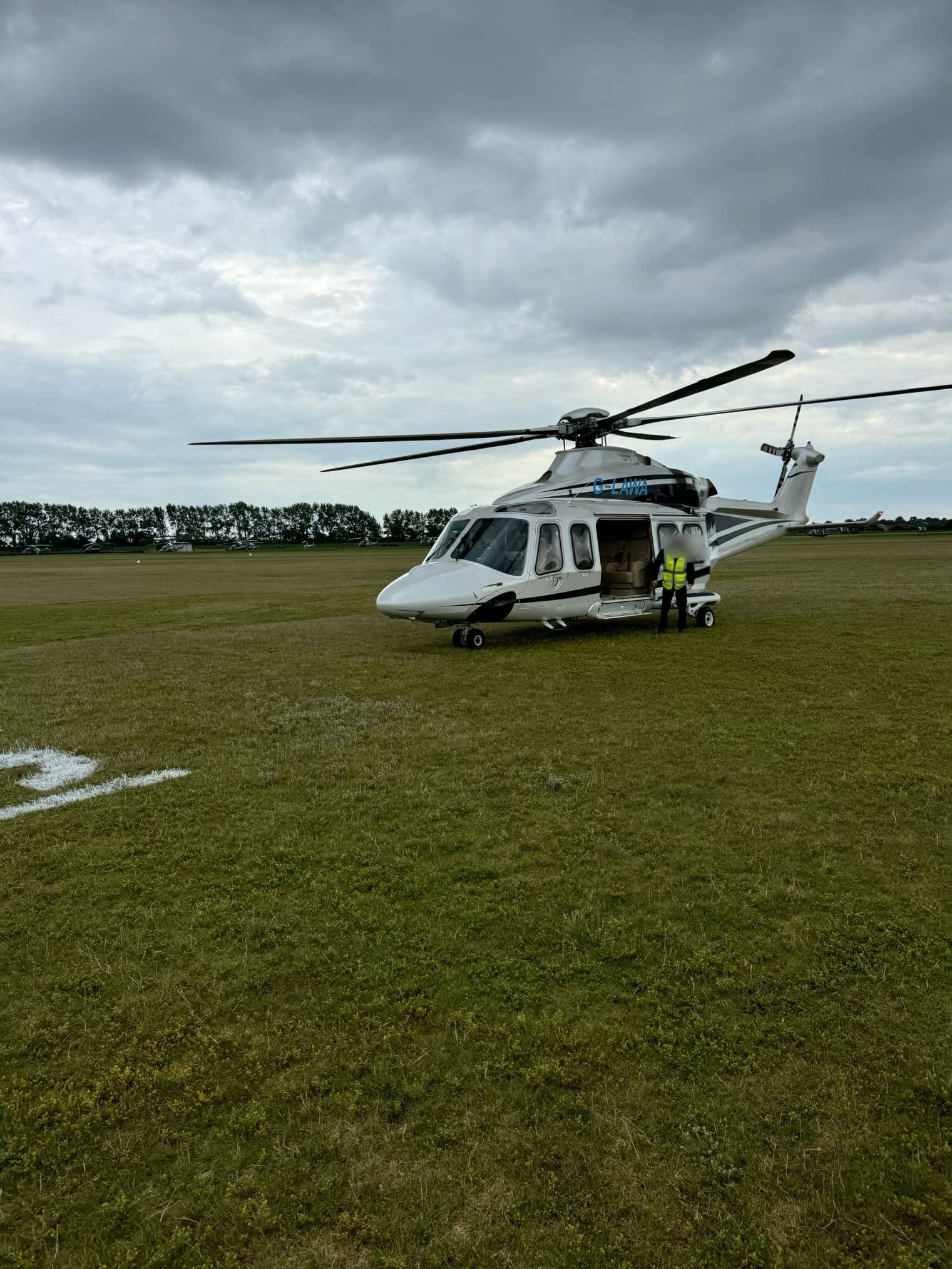 A white helicopter sits on a grassy airfield under a cloudy sky with crew members near the open door.