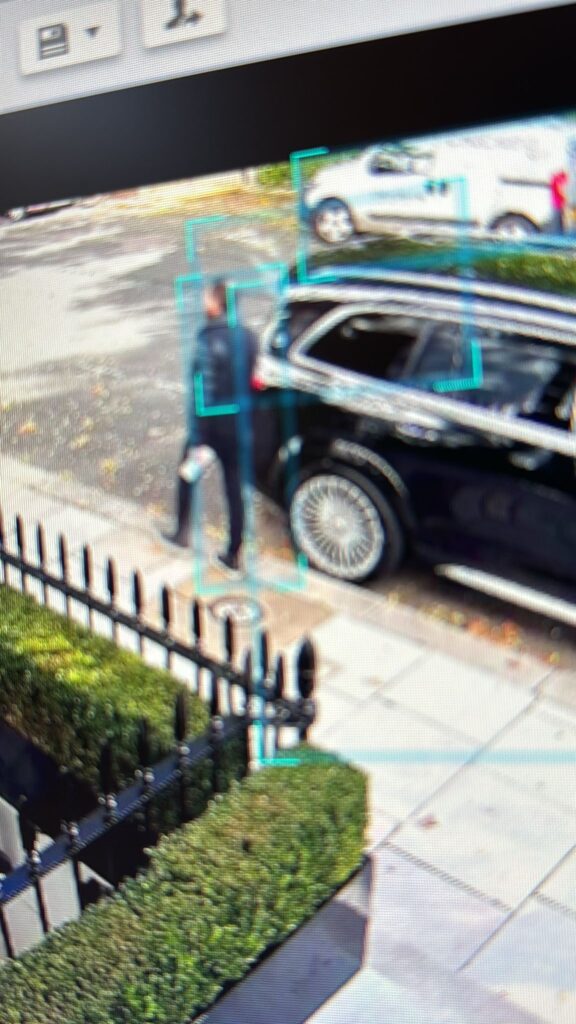 A CCTV screen captures a person standing beside a black vehicle near a street curb.