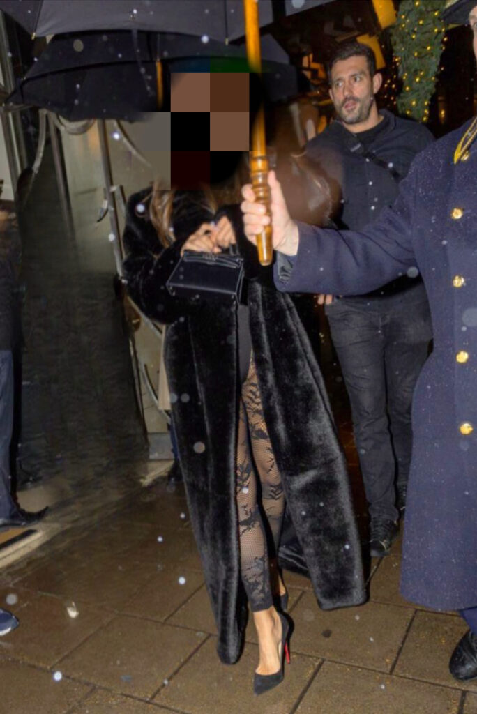 A woman dressed in a long black fur coat and heels walks under an umbrella held by someone else on a rainy night.